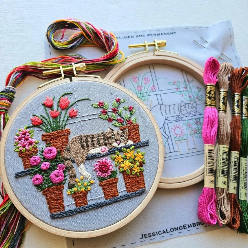 The Jessica Long Embroidery 5 Cat Nap with Flowers Embroidery Kit features a tabby cat sleeping on shelves with potted flowers, colorful threads, and an extra hoop with the outlined design—ideal for beginner embroiderers.