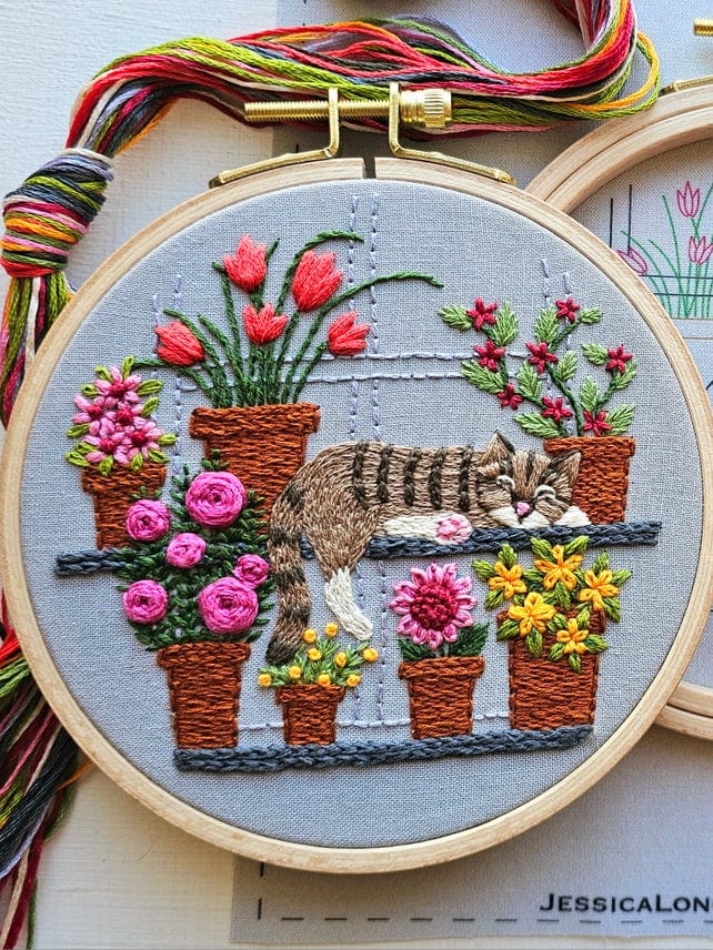 The 5 Cat Nap with Flowers Embroidery Kit by Jessica Long Embroidery features a detailed tabby cat sleeping on a shelf, surrounded by tulips, roses, and yellow blooms, with embroidery threads included.