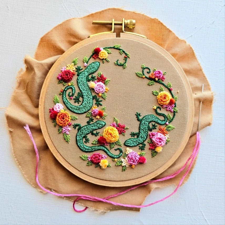 The Jessica Long Embroidery 6 Beginner Backyard Geckos Embroidery Kit features tan fabric in a hoop, embroidered with colorful geckos and vibrant flowers. Ideal for beginners, the kit includes thread extending from the hoop.
