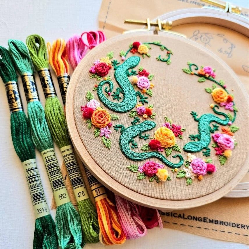 The 6 Beginner Backyard Geckos Embroidery Kit by Jessica Long Embroidery features green, ornate S shapes with colorful flowers. Included are skeins of green, yellow, orange, and pink floss. A faint pattern appears on fabric in another hoop.