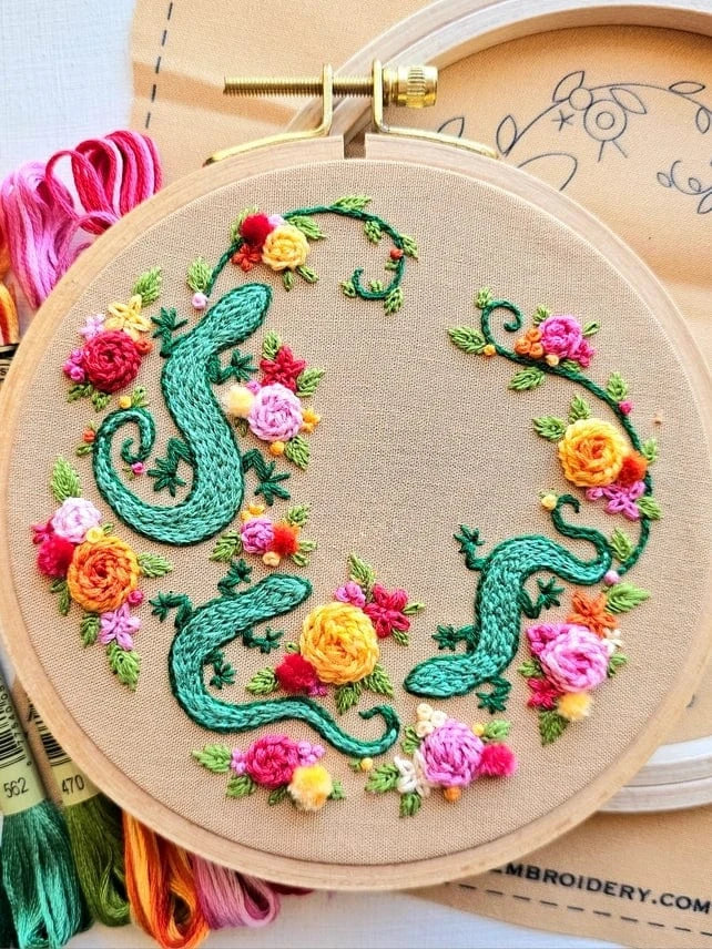 The 6 Beginner Backyard Geckos Embroidery Kit by Jessica Long Embroidery features two green geckos among colorful flowers on tan fabric, perfect for beginners. The kit, pattern, and threads are also shown in the background.