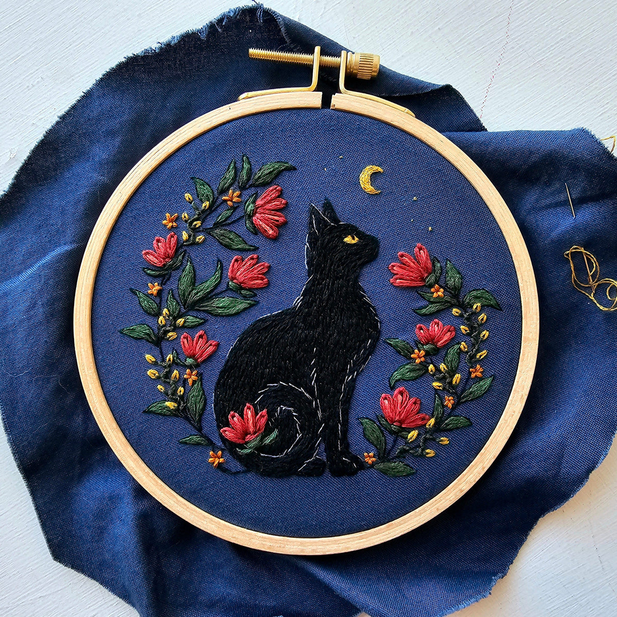 The 5 Beginner Stargazer Black Cat Embroidery Kit by Jessica Long Embroidery features a black cat gazing at a yellow crescent moon, surrounded by red flowers and green leaves on dark blue fabric in a wooden hoop.