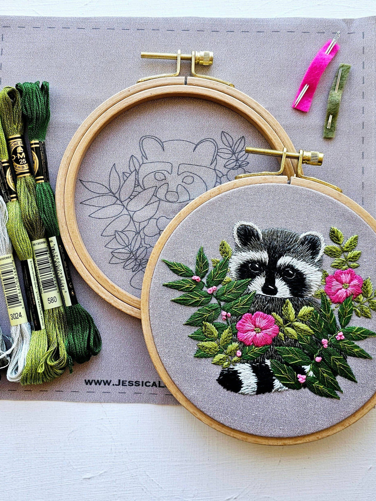 The 5 Raccoon Embroidery Kit by Jessica Long Embroidery includes a finished, outlined hoop, green threads, two needles, and a pink threader on grey fabric—ideal for practicing thread painting with a raccoon and pink flowers design.