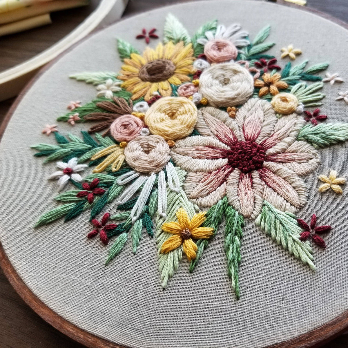 The 6 Beginner Floral Harvest Embroidery Kit by Jessica Long Embroidery features hand-stitched sunflowers, daisies, and leaves in yellow, cream, green, and burgundy on beige fabric—ideal for adding unique charm to your home decor.