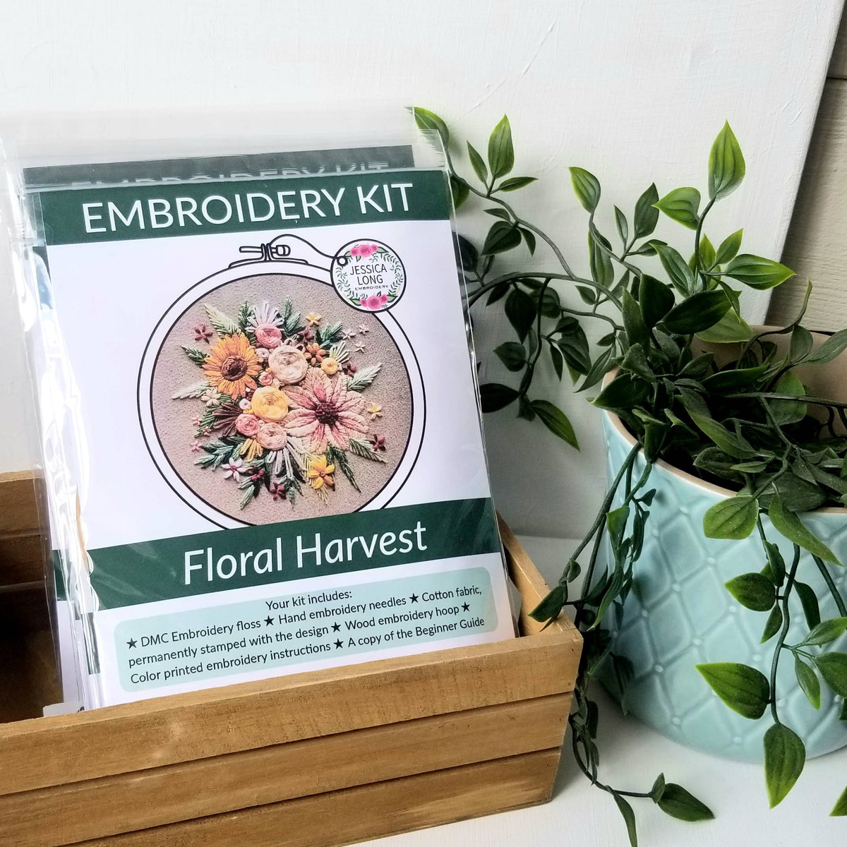 A 6 Beginner Floral Harvest Embroidery Kit by Jessica Long Embroidery sits in a wooden box beside a green potted plant. The packaging displays hand-stitched flowers, ideal for home decor or creative projects.