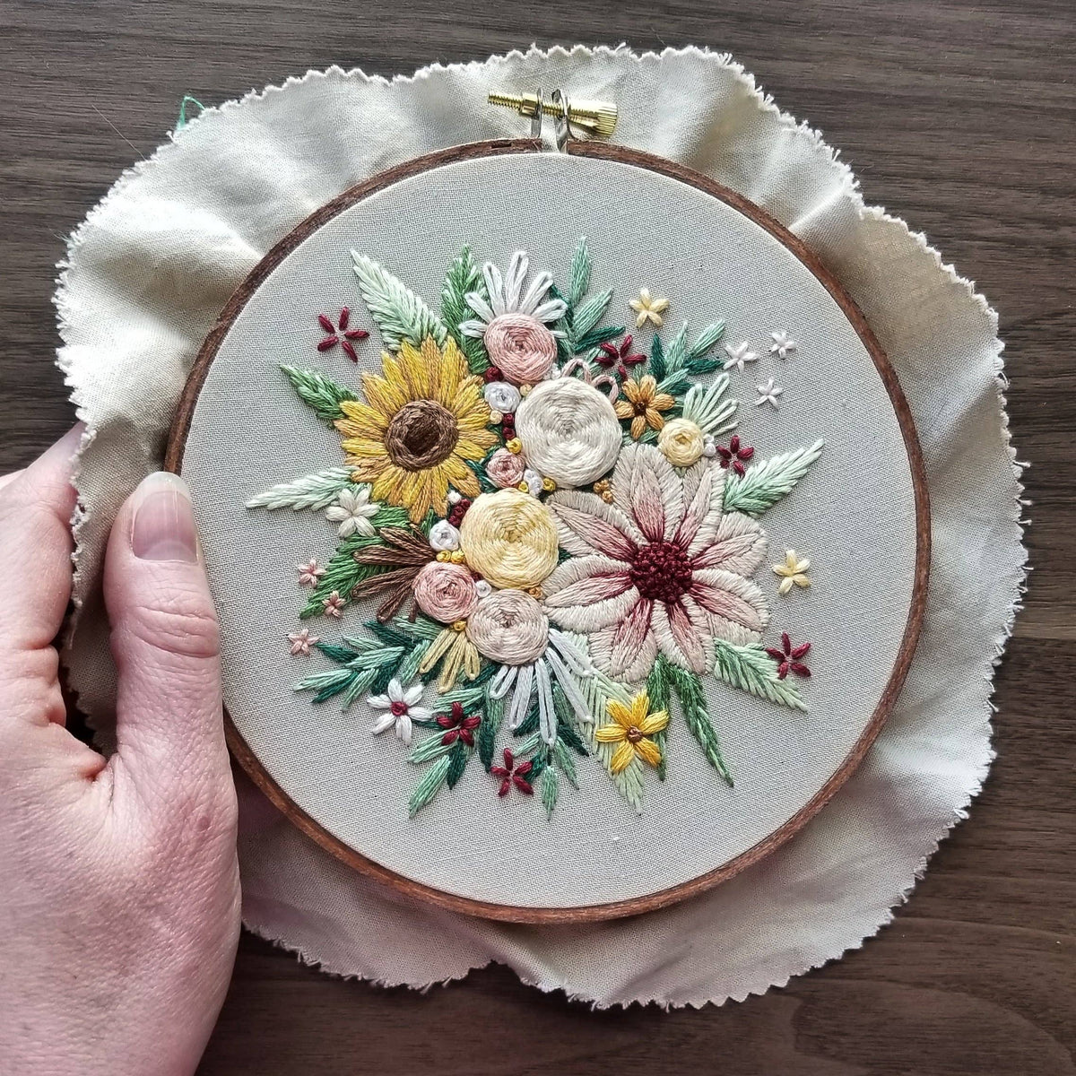 A hand holds the Jessica Long Embroidery 6 Beginner Floral Harvest Embroidery Kit—featuring hand-stitched sunflowers, roses, and more on beige fabric—creating beautiful home decor against a wooden surface.