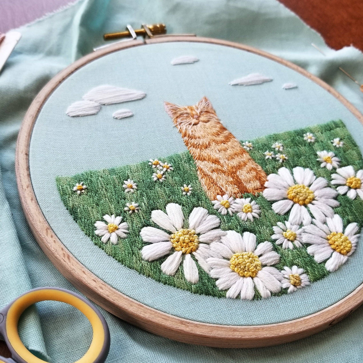 The 5 Happy Cat Embroidery Kit by Jessica Long Embroidery features an orange cat in a daisy field under a cloudy blue sky. Ideal as a hand sewing project or beginner embroidery kit, shown with scissors on light blue fabric.