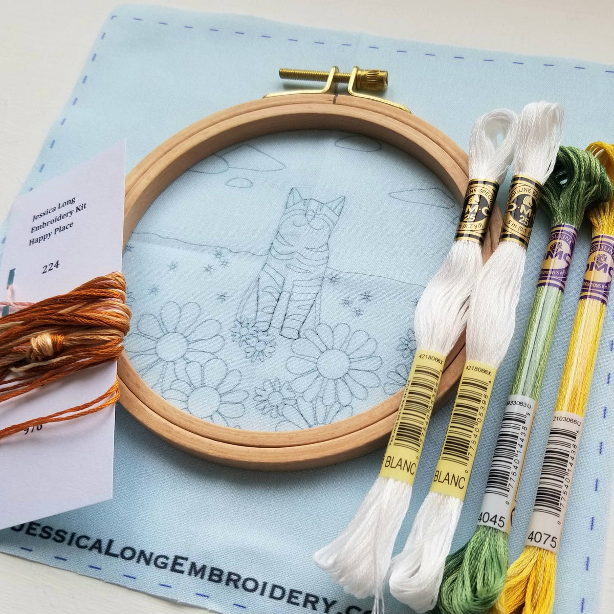The 5 Happy Cat Embroidery Kit by Jessica Long Embroidery is ideal for beginner embroidery, featuring cat and flower outlines in a wooden hoop, with colorful flosses and a thread card displayed on blue cloth.