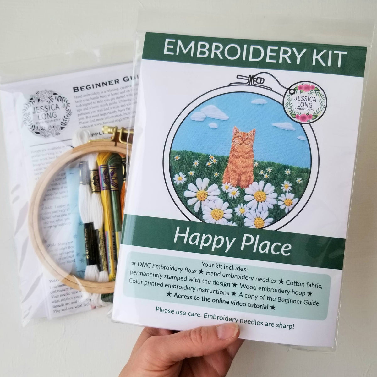 A hand holds two Jessica Long Embroidery kits—a 5 Happy Cat Embroidery Kit featuring an orange cat in daisies called Happy Place, and a second kit with hoops, floss, and a guide—great for beginner embroidery or hand sewing projects.