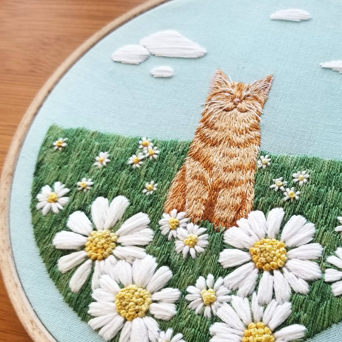 The 5 Happy Cat Embroidery Kit by Jessica Long Embroidery is a beginner-friendly set featuring an orange cat among white daisies in a green field under a blue sky, all framed in a classic wooden hoop.