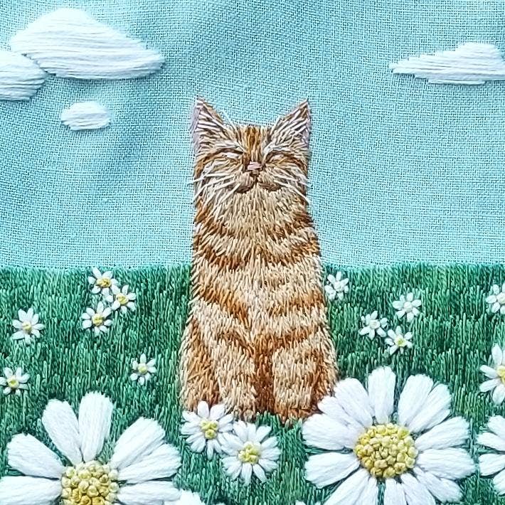 The 5 Happy Cat Embroidery Kit by Jessica Long Embroidery features a fluffy orange cat in a green field with daisies and embroidered clouds—a charming hand sewing project suitable for all skill levels.