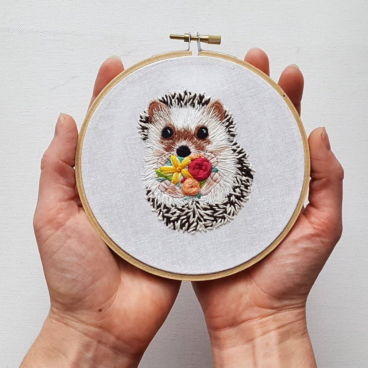 A pair of hands holds the Jessica Long Embroidery 6 Hedgehog Embroidery Kit, featuring detailed hedgehog and colorful flower stitching on white fabric—an ideal choice for beginner embroidery enthusiasts.