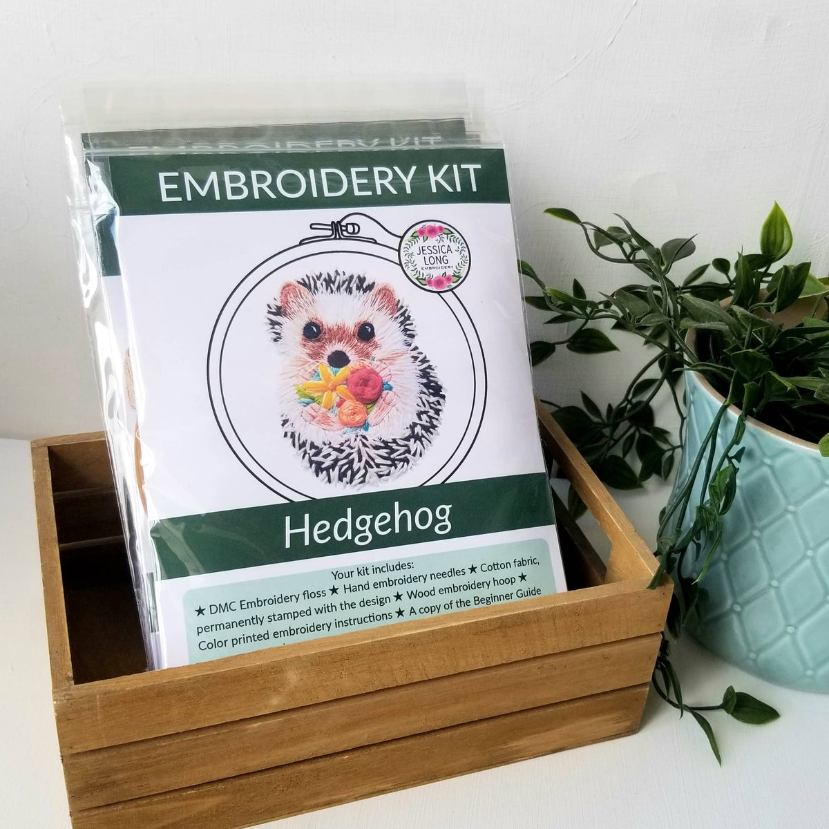 The Jessica Long Embroidery 6 Hedgehog Embroidery Kit includes floss, needle, fabric, and instructions for beginners, featuring hoop art of a hedgehog holding fruit—all in a charming wooden box by a potted green plant.