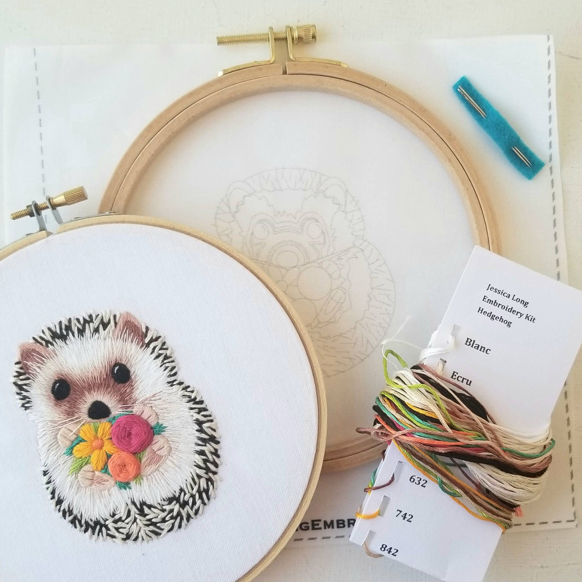 The Jessica Long Embroidery 6 Hedgehog Embroidery Kit includes a finished hoop with a hedgehog holding flowers, an empty hoop with the pattern, colorful threads, and a blue needle threader—perfect for beginners.