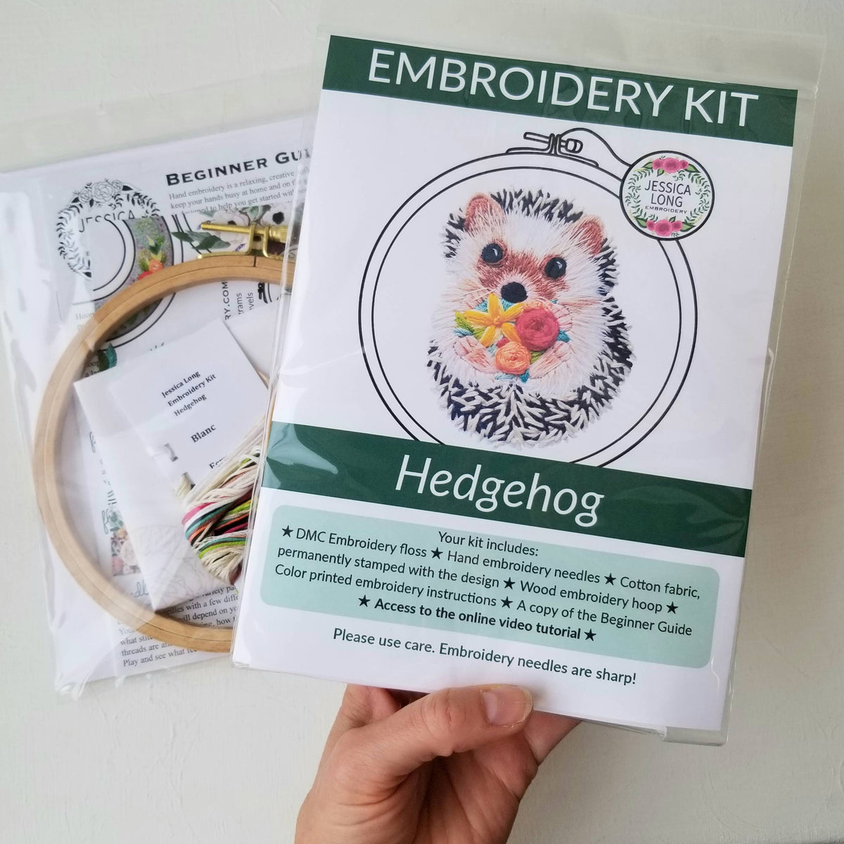 A hand holds the Jessica Long Embroidery 6 Hedgehog Embroidery Kit, ideal for beginners. The kit includes fabric, floss, a wooden hoop, needles, instructions, and a beginner guide. Another packaged kit is visible in the background.