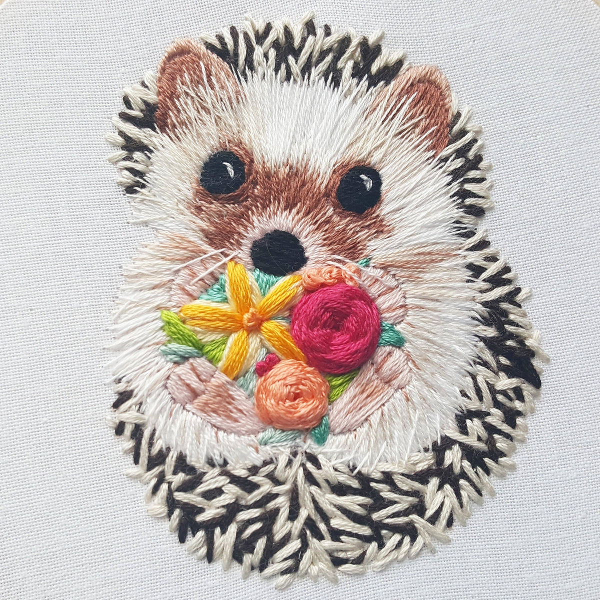 The 6 Hedgehog Embroidery Kit by Jessica Long Embroidery features a cute brown and cream hedgehog holding colorful flowers with green leaves, stitched on white fabric—perfect for hoop art or beginner embroidery projects.