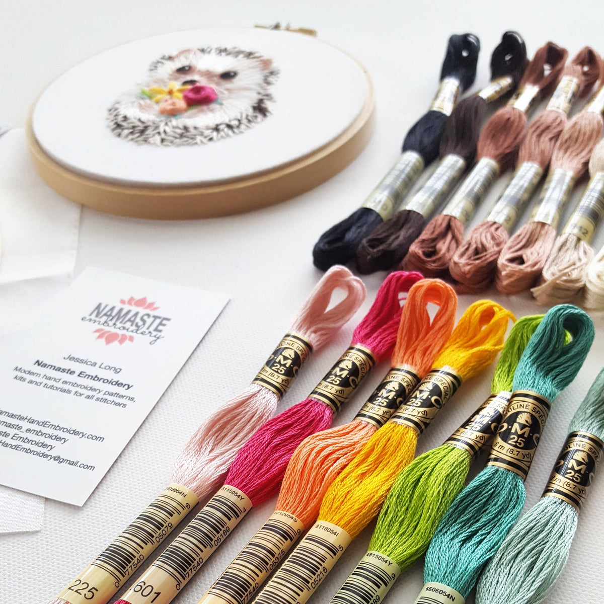 The 6 Hedgehog Embroidery Kit by Jessica Long Embroidery is displayed with colorful floss, finished hoop art, and a business card on a white surface, highlighting materials and branding for beginner embroidery projects.