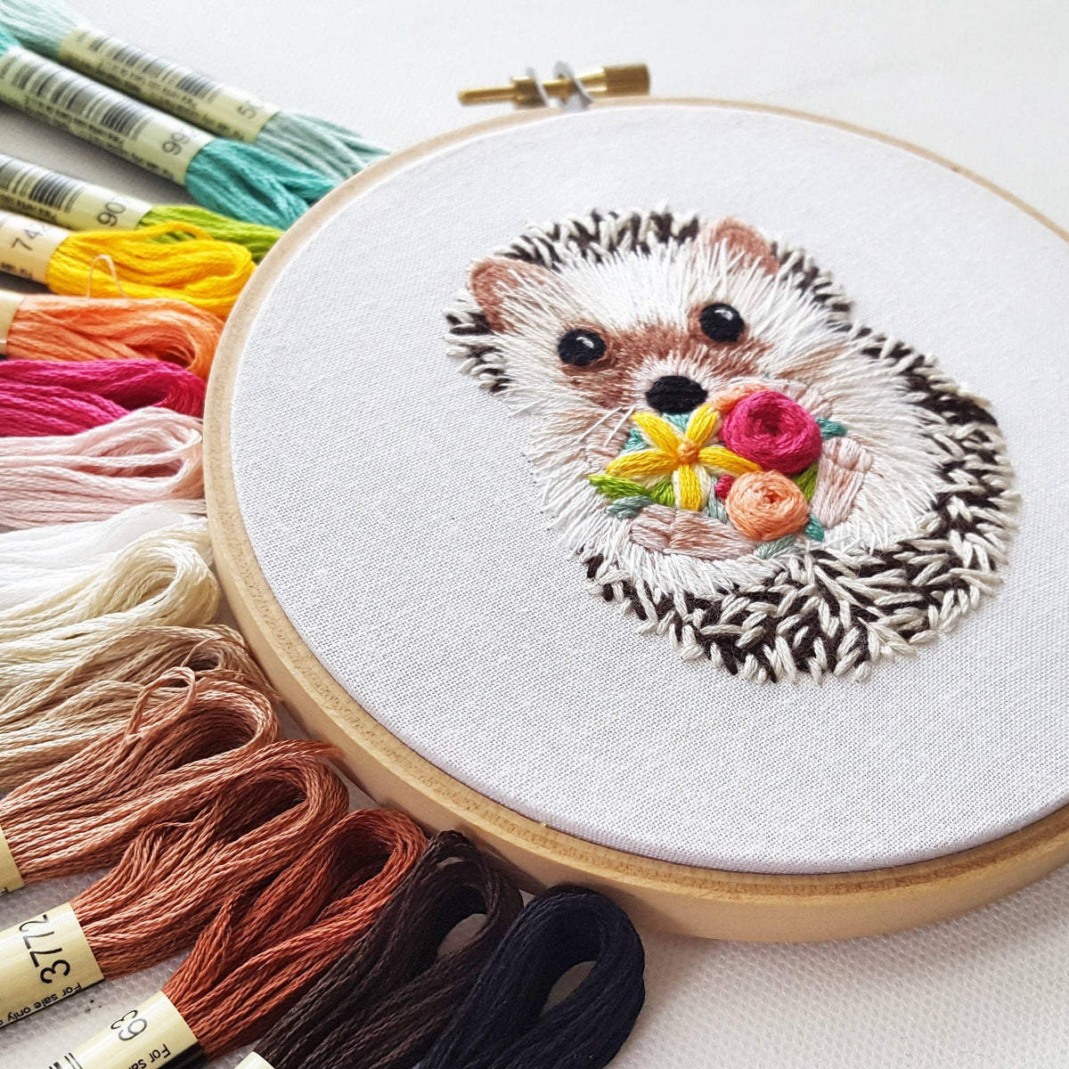 The 6 Hedgehog Embroidery Kit by Jessica Long Embroidery features a stitched hedgehog with colorful flowers, surrounded by assorted embroidery floss—an ideal beginner embroidery or hedgehog-themed kit.
