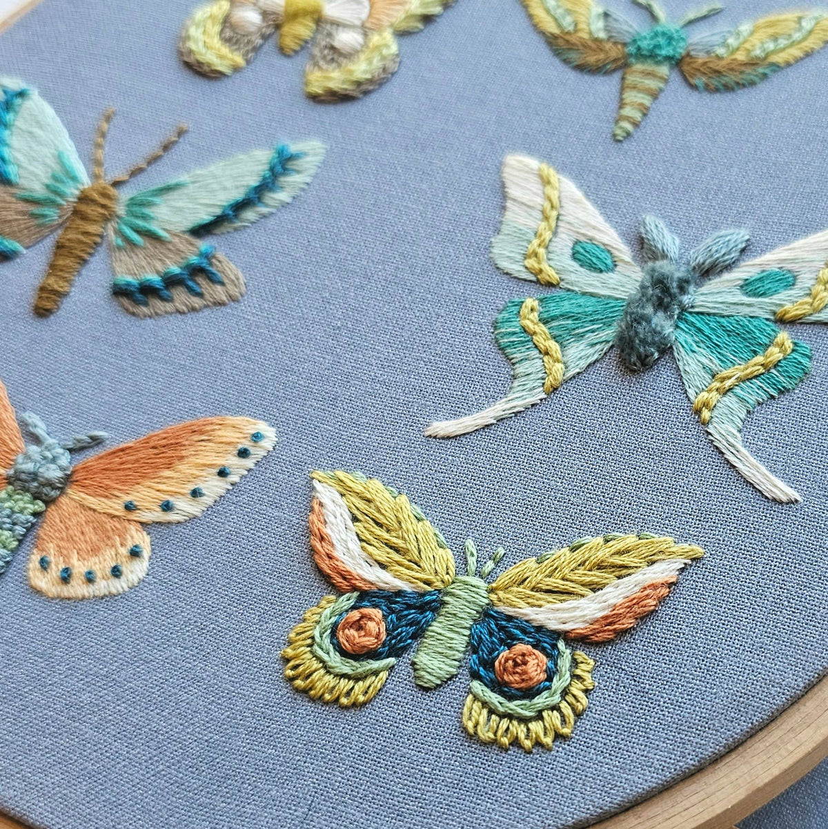 The *Late May Pre-Order* 6 Moth Embroidery Kit by Jessica Long Embroidery features vibrant moths and butterflies on light blue fabric—ideal for hand embroidery fans or as a unique addition to your kit.