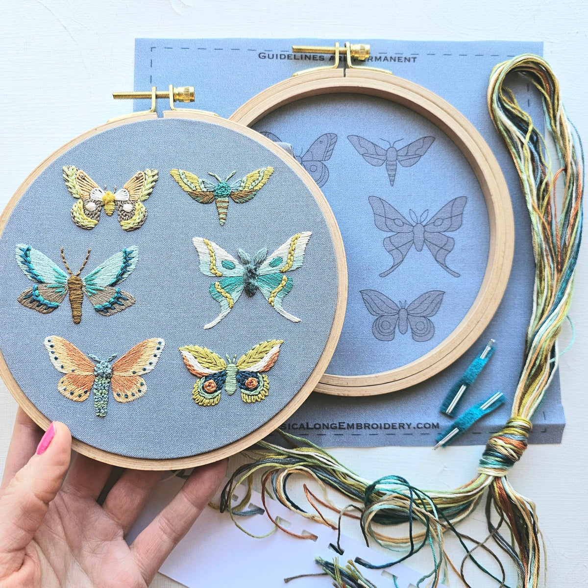 A hand holds a *Late May Pre-Order* 6 Moth Embroidery Kit by Jessica Long Embroidery, showing six colorful moths on gray fabric. Nearby are an empty hoop, embroidery floss, needles, and a kit pattern sheet with moth outlines.