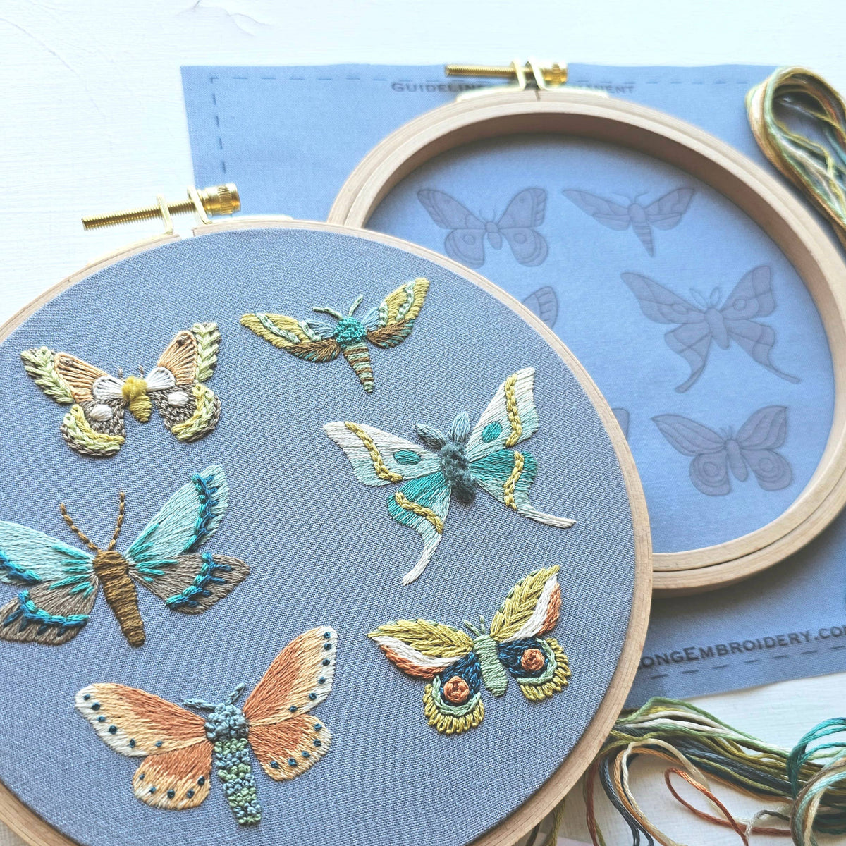 A detailed, colorful moth embroidery is displayed in a wooden hoop, with the Jessica Long Embroidery *Late May Pre-Order* 6 Moth Embroidery Kit—including printed pattern and threads—resting nearby on a white surface.