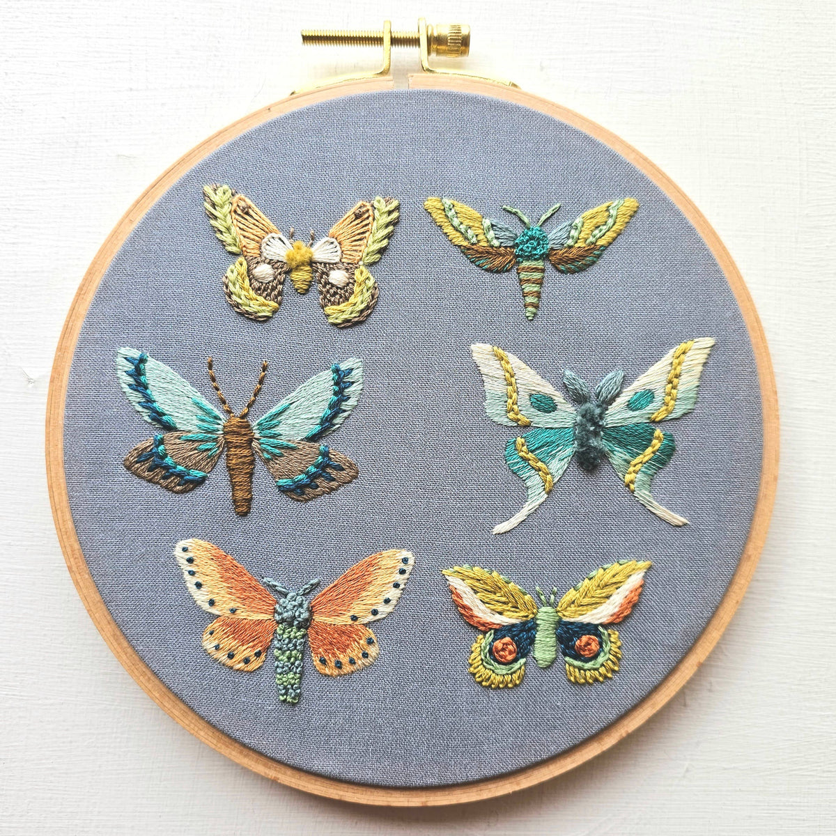 The *Late May Pre-Order* 6 Moth Embroidery Kit by Jessica Long Embroidery lets you stitch seven unique jeweled moths on gray fabric, with a step-by-step video tutorial—perfect for hand embroidery fans.