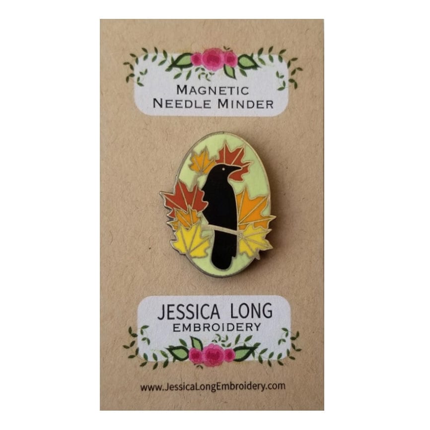The Autumn Birds Enamel Needle Minder by Jessica Long Embroidery features a black crow among autumn leaves, displayed on a branded card.