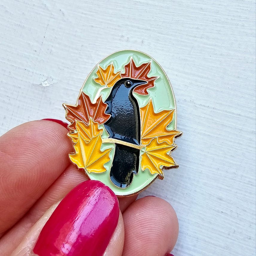 A hand with bright pink nail polish holds the Autumn Birds Enamel Needle Minder by Jessica Long Embroidery, featuring a black crow among orange and yellow autumn leaves on a pale background.