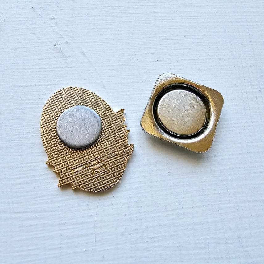 Two gold-colored metal pin backs—a circular one attached to the Autumn Birds Enamel Needle Minder by Jessica Long Embroidery, and a square one—rest on a white textured surface.