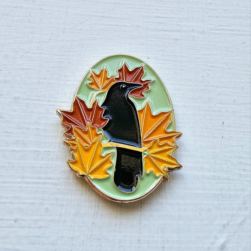 The Autumn Birds Enamel Needle Minder by Jessica Long Embroidery features a black crow with red, orange, and yellow maple leaves on a pale green oval background, shown resting on a textured white surface.