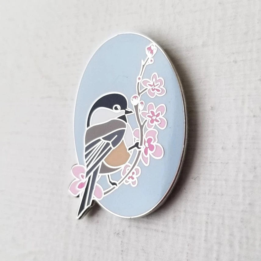 The Chickadee Enamel Needle Minder by Jessica Long Embroidery features a stylish black-capped chickadee on a branch with pink flowers, set on a blue oval. Perfect as both an elegant enamel pin and a charming magnetic needle minder for crafts.