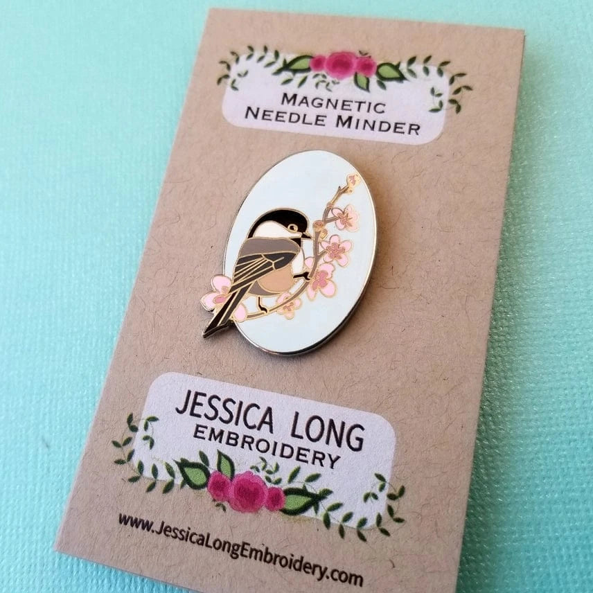 The Chickadee Enamel Needle Minder by Jessica Long Embroidery features a bird on a flowering branch and comes attached to a floral card with a light blue textured background.