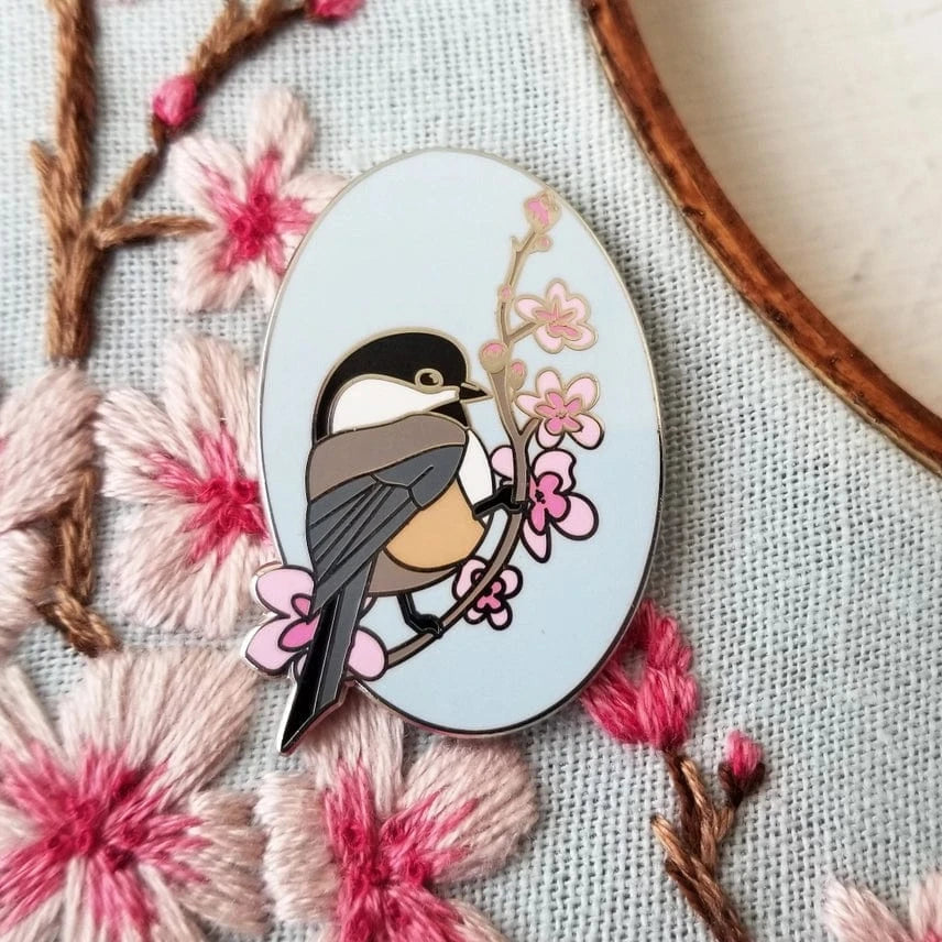 The Chickadee Enamel Needle Minder by Jessica Long Embroidery features a magnetic hard enamel pin of a chickadee on a flowering branch, set on fabric with embroidered cherry blossoms—ideal for keeping your needle in place.