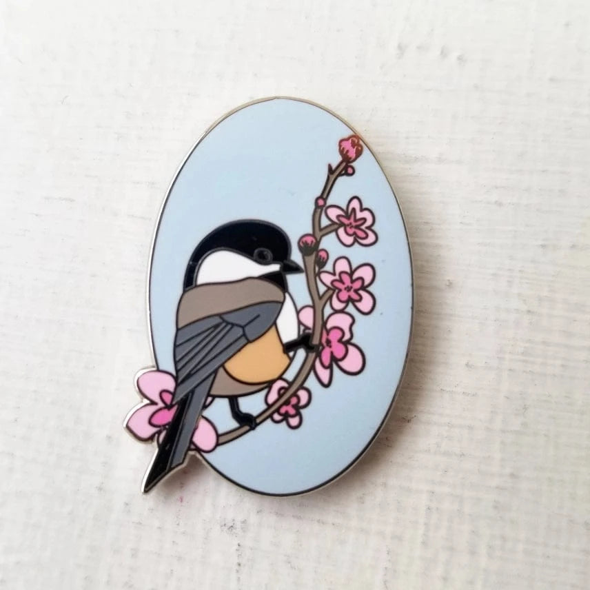 The Chickadee Enamel Needle Minder by Jessica Long Embroidery features a stylized chickadee on a blossom branch with a pale blue background—perfect as a magnetic pin or needle minder for your crafting projects.