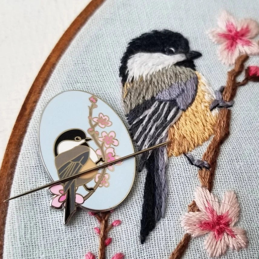 The Chickadee Enamel Needle Minder by Jessica Long Embroidery is a hard enamel pin with a black-capped chickadee and cherry blossoms, shown resting on an embroidered hoop—perfect for holding your needle while you stitch.
