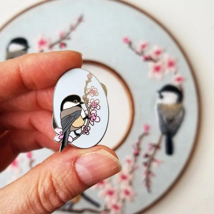 A hand holds the Jessica Long Embroidery Chickadee Enamel Needle Minder, an oval hard enamel pin featuring a bird on a cherry blossom branch; a similar floral design appears softly blurred in the background.
