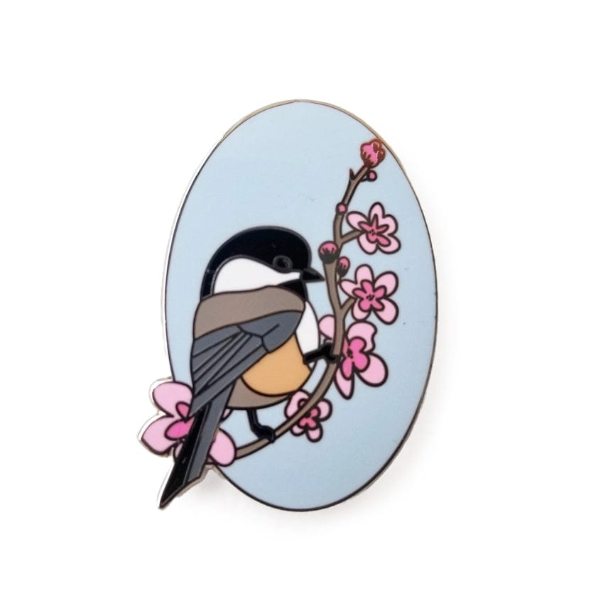 The Chickadee Enamel Needle Minder by Jessica Long Embroidery features a stylized chickadee on a branch with pink flowers and a light blue background; great as a magnetic pin or decorative needle minder.