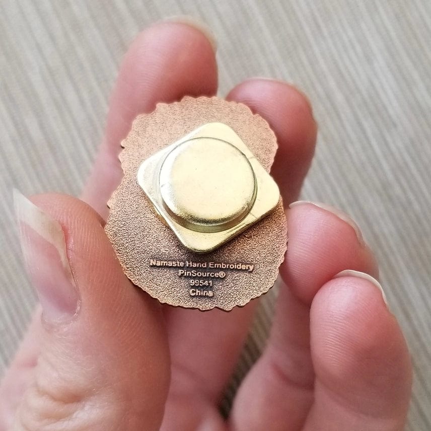 A hand holds the back of a Hedgehog Enamel Needle Minder by Jessica Long Embroidery with a gold clasp, shown against a textured tan background.