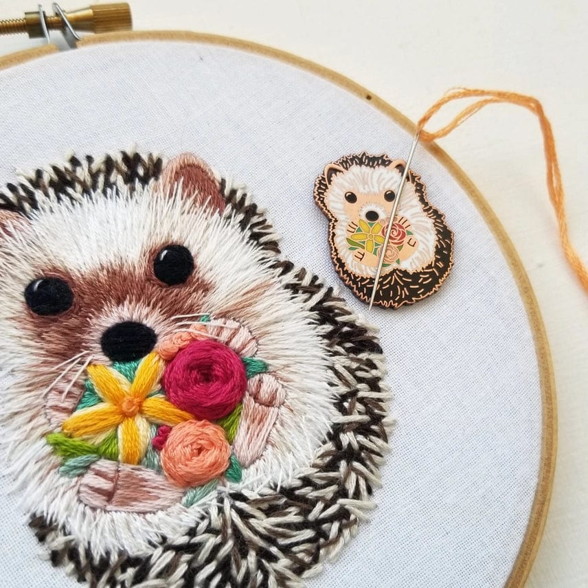 A hoop displays hedgehog embroidery with colorful flowers. Beside it, the Hedgehog Enamel Needle Minder by Jessica Long Embroidery holds an orange thread and needle, adding charm and function as a magnetic pin.