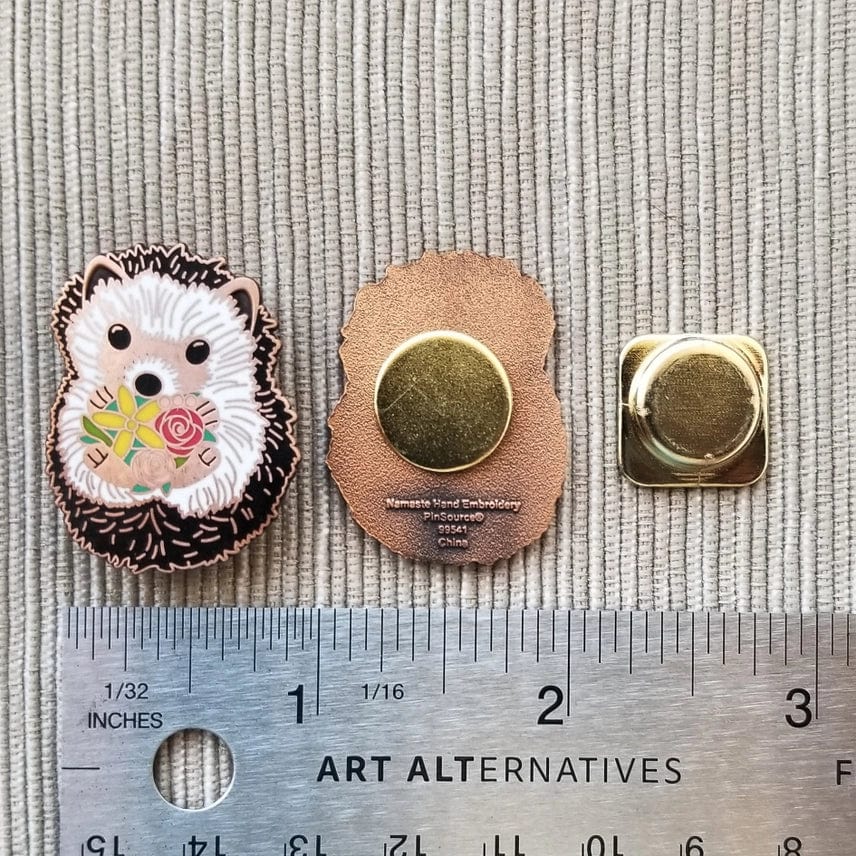 The Hedgehog Enamel Needle Minder by Jessica Long Embroidery features a floral hedgehog design. Shown front and back with its metal clutch, it measures about 1.25 inches tall, displayed on textured fabric with a ruler for scale.