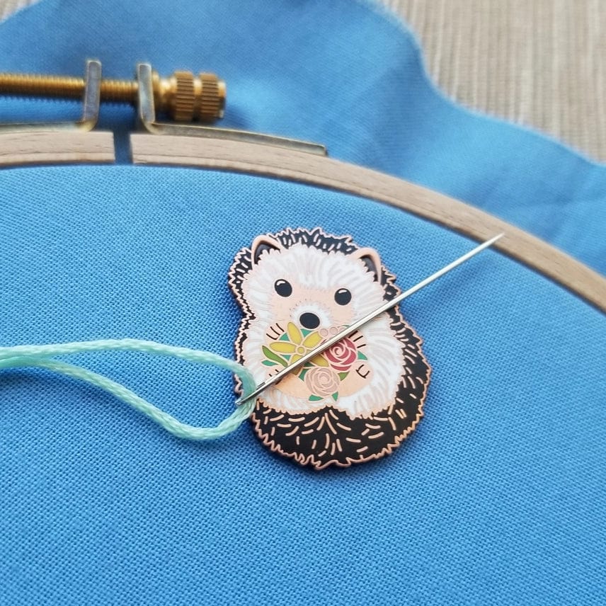 A light blue threaded needle passes through the Hedgehog Enamel Needle Minder by Jessica Long Embroidery, which features a floral design and rests on blue fabric in an embroidery hoop.