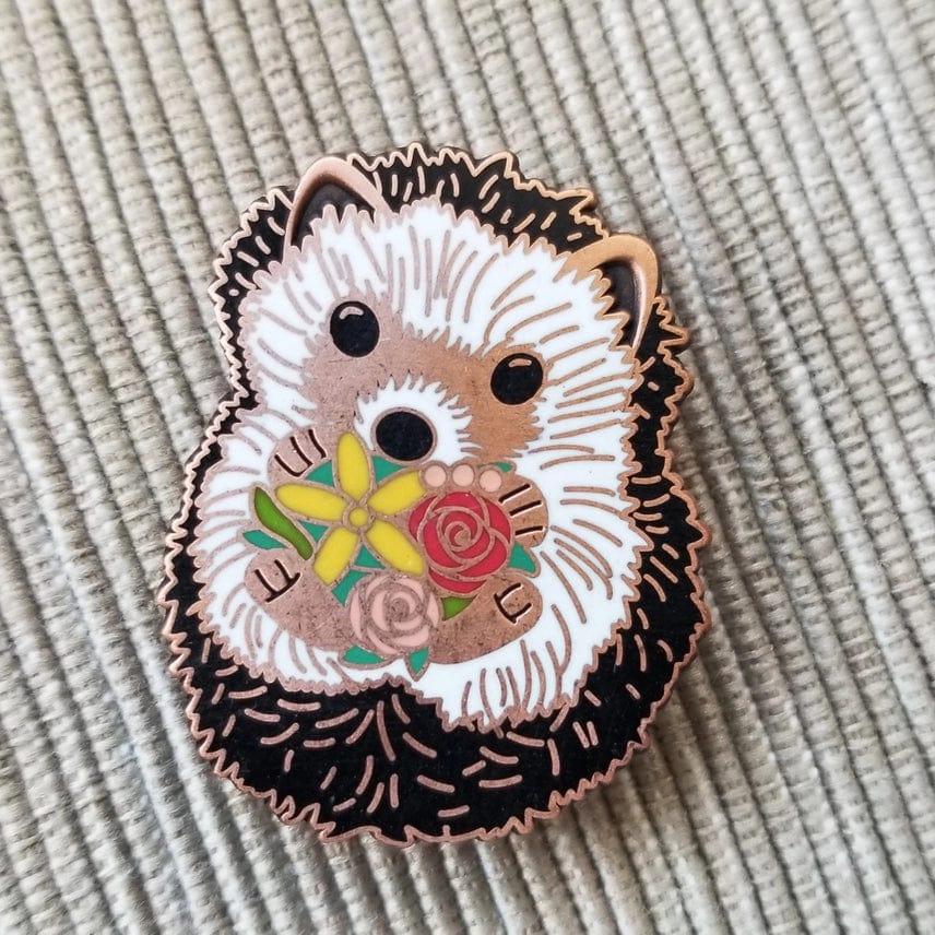 The Hedgehog Enamel Needle Minder by Jessica Long Embroidery is a cute hard enamel pin featuring a hedgehog holding colorful flowers, including yellow and red roses. This magnetic pin keeps needles secure and adds charm to your workspace.
