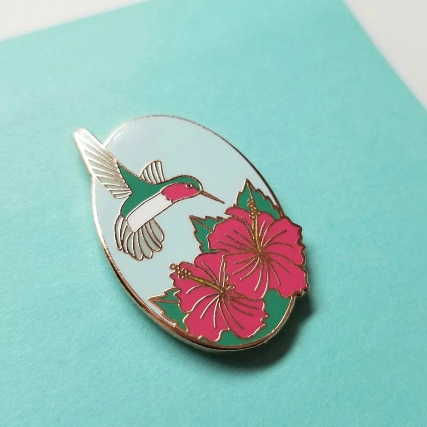 The Hummingbird Enamel Needle Minder by Jessica Long Embroidery features a colorful hummingbird with pink hibiscus flowers on a light blue oval, making it a perfect magnetic pin for needlework enthusiasts.