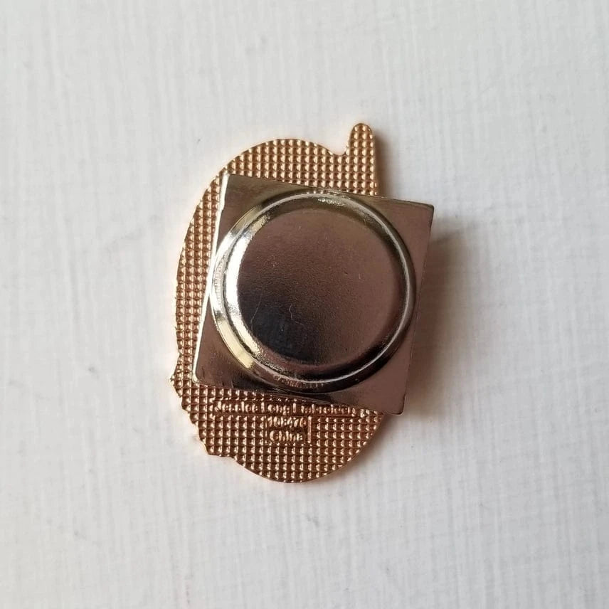 A close-up of the back of the Hummingbird Enamel Needle Minder by Jessica Long Embroidery, showing its gold hard enamel finish and a round silver butterfly clutch fastener on a white textured surface.