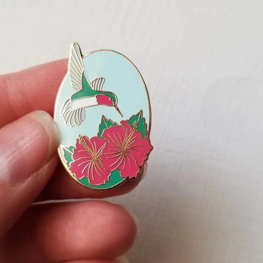 A hand displays the Hummingbird Enamel Needle Minder by Jessica Long Embroidery, featuring a green and white hummingbird above pink hibiscus flowers with green leaves on a light blue background. Ideal for stylish, magnetic needle keeping.