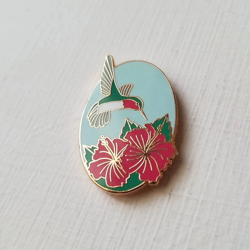 The Hummingbird Enamel Needle Minder by Jessica Long Embroidery features a hummingbird in flight above two red hibiscus flowers with green leaves on a pale blue oval—perfect as a magnetic accessory for your sewing projects.