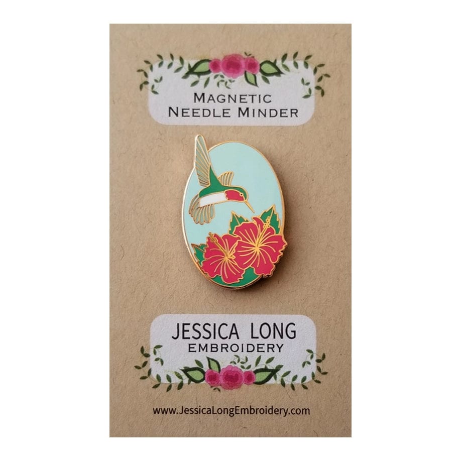The Hummingbird Enamel Needle Minder by Jessica Long Embroidery features pink flowers and is displayed on a floral card marked “MAGNETIC NEEDLE MINDER.” It’s perfect for securely holding your magnetic pin during stitching.