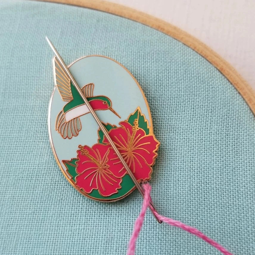 The Jessica Long Embroidery Hummingbird Enamel Needle Minder, featuring red hibiscus flowers, magnetically holds your needle and pink thread on blue fabric in a wooden hoop—keeping your stitching secure and stylish.