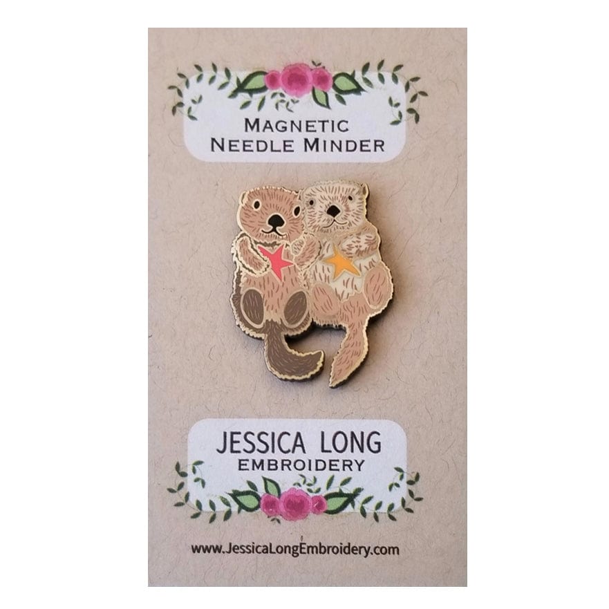 The Otterly Adorable Enamel Needle Minder by Jessica Long Embroidery features two cuddling otters with starfish. This charming magnetic pin comes on a floral backing card—perfect for your stitching projects.