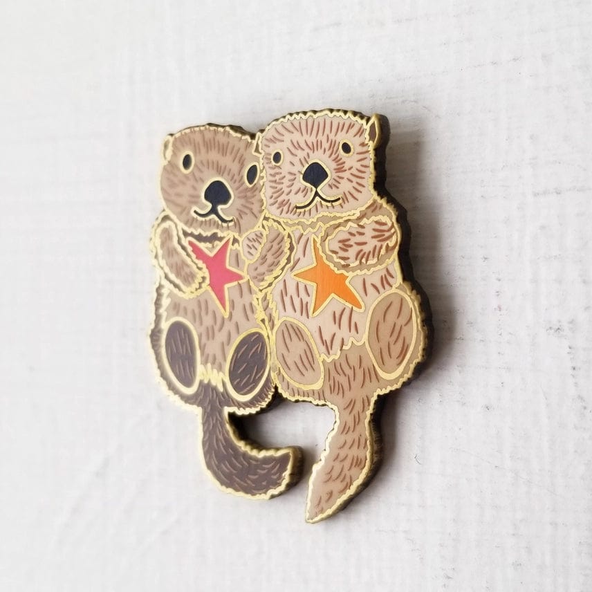 The Otterly Adorable Enamel Needle Minder by Jessica Long Embroidery features two illustrated sea otters with orange and red starfish on a light textured background. Perfect for keeping your needles in place while crafting.
